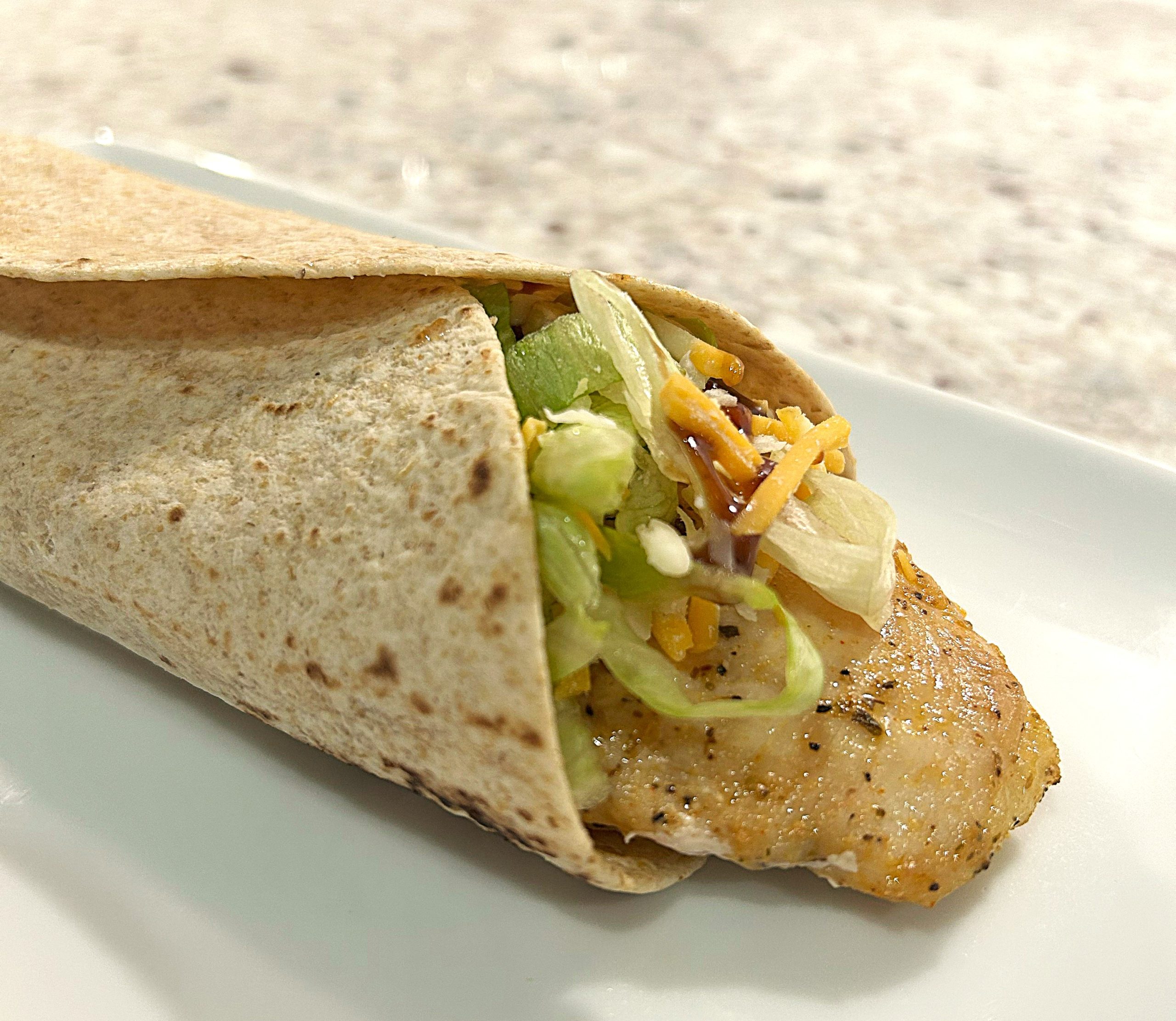 A piece of chicken wrapped in a whole wheat tortilla with shredded lettuce, shredded cheese and barbeques sauce.