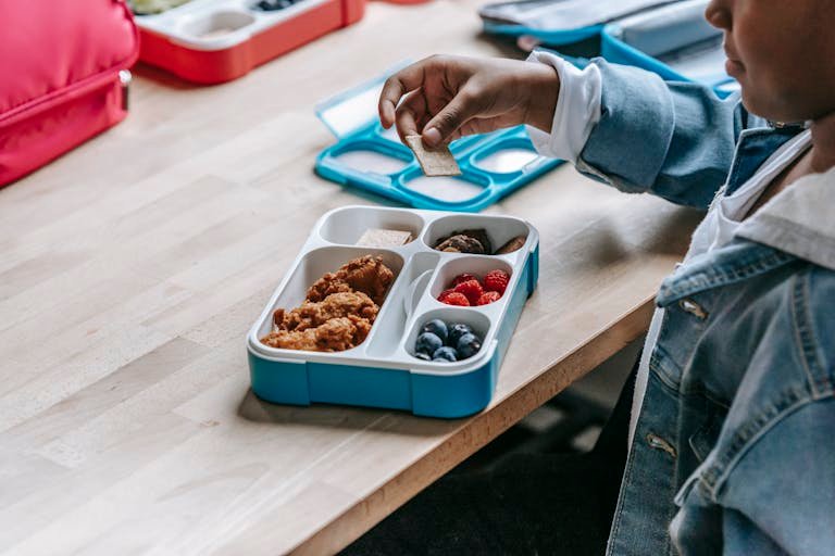 From above side view of crop unrecognizable ethnic school kid at table with lunch container full of tasty food for back to school