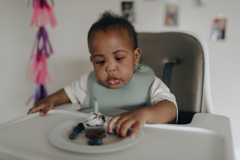Adorable baby in high chair using baby led weaning to savor a cupcake with a candle. Celebratory mood indoors.
